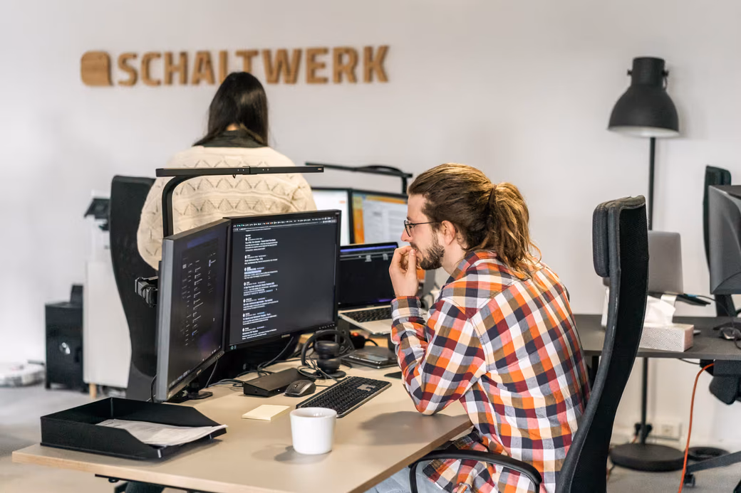 Ein Entwickler sitz an seinem Arbeitsplatz am Schreibtisch und arbeitet konzentriert. Im Hintergrund sieht man eine weitere Mitarbeiterin.