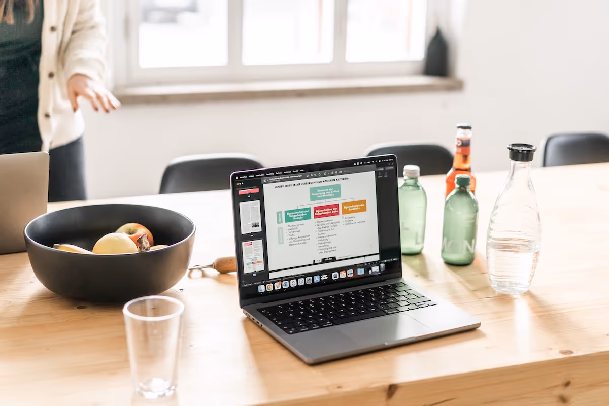 Laptop displaying a colorful chart on a wooden table with fruit bowl, several bottles, and a clear glass cup.