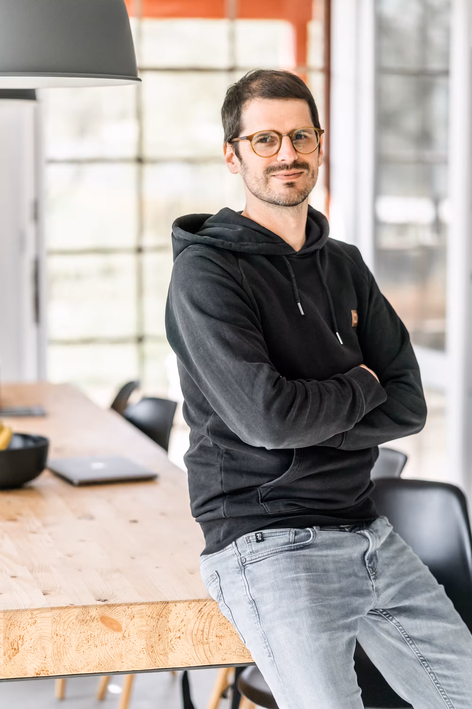 Tobi steht mit verschränkten Armen lächelnd in einem modernen Büro mit Glasfront.